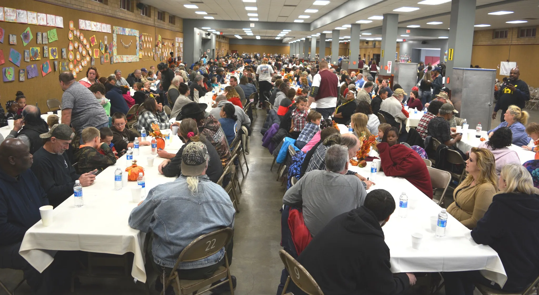 A busy dining hall with neighbors sharing a meal at long tables.
