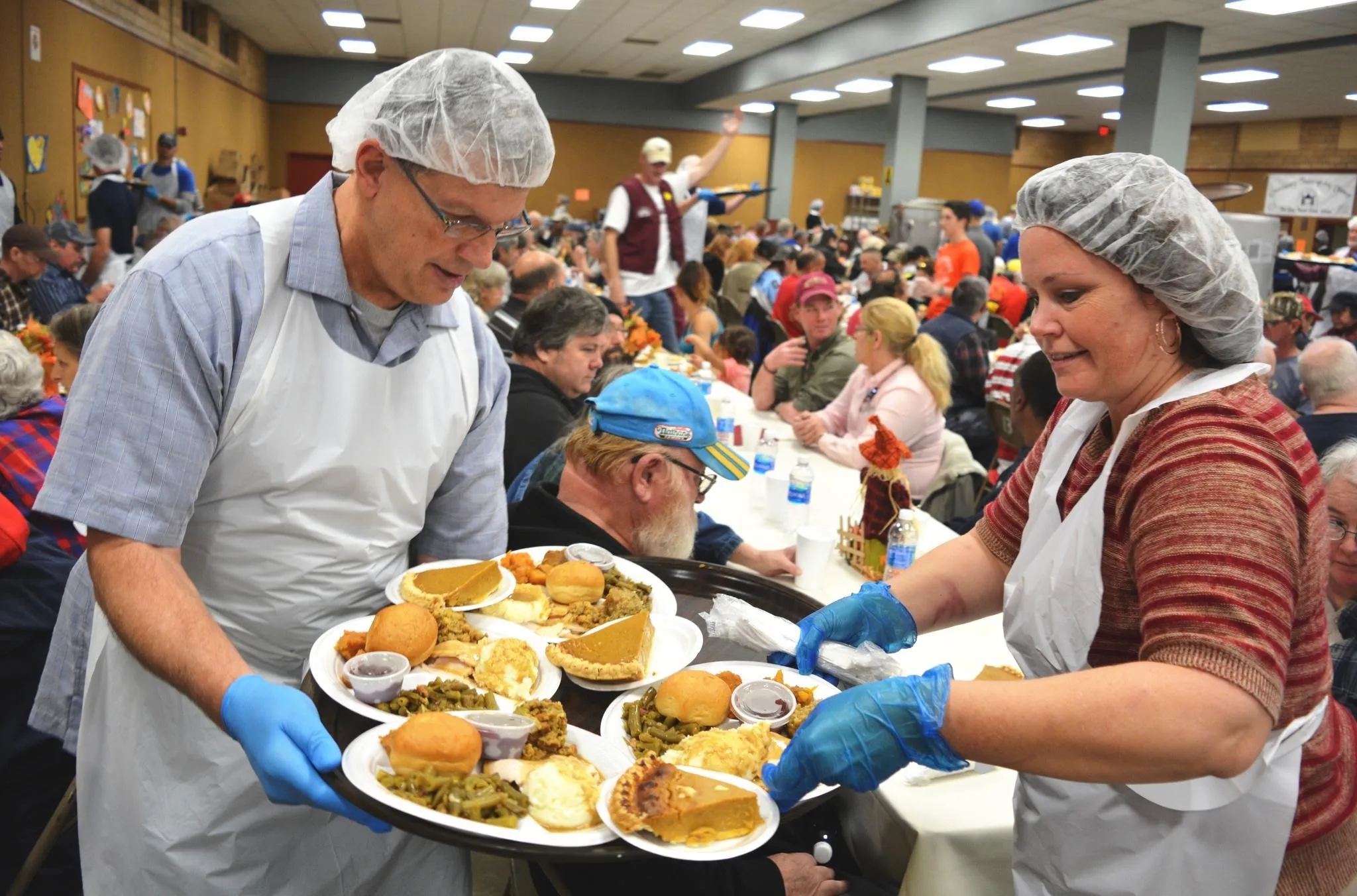A man in blue holds a tray of Thanksgiving dinners while a woman in red stripes picks up plates.